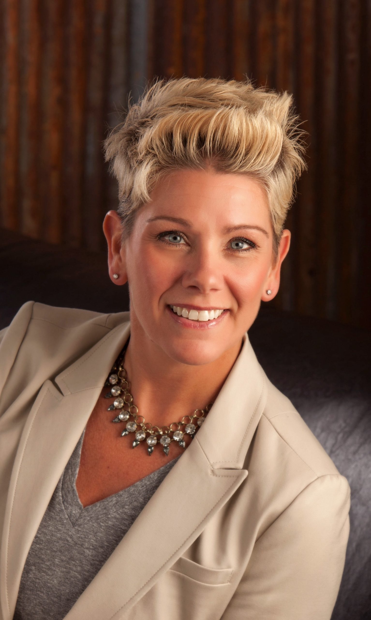 Smiling woman with short bleached-blonde hair, wearing a beige blazer and layered necklace, against a dark wooden background.