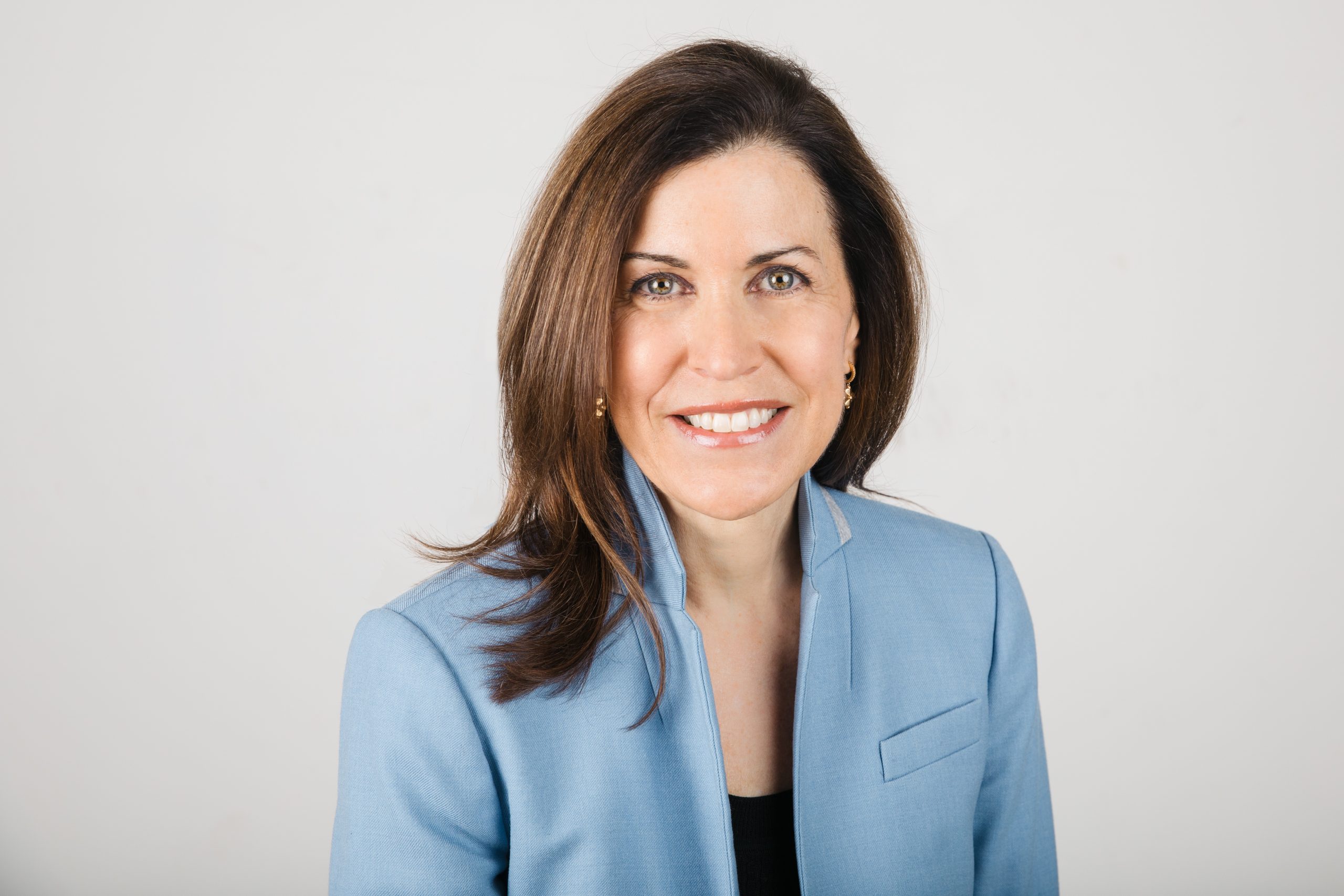 Smiling woman with shoulder-length brown hair in a light blue blazer, neutral background.